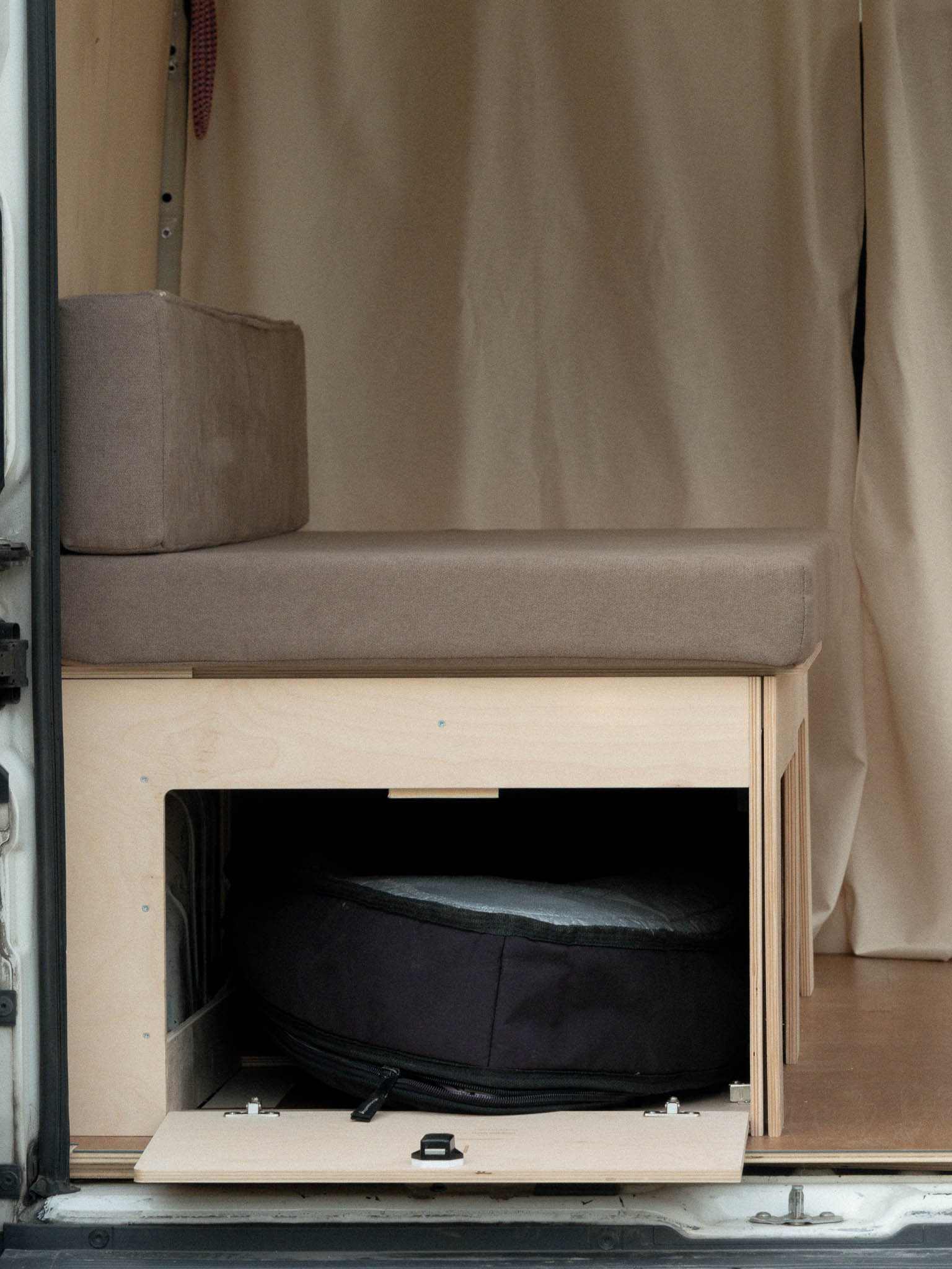 Letto Nom Outdoor in modalità divano con anta laterale aperta e spazio di stivaggio per tavola da surf, soluzione modulare per camperizzazione van ideale per surfisti.