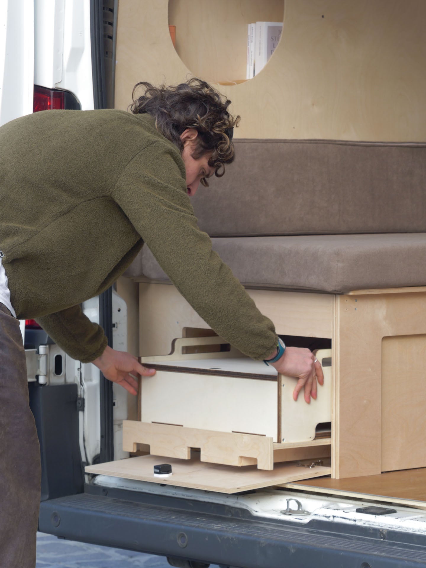 Persona che inserisce il Totem Cook nel piano estraibile laterale del Letto Nom Outdoor, sistema modulare per camperizzazione van con cucina integrabile.